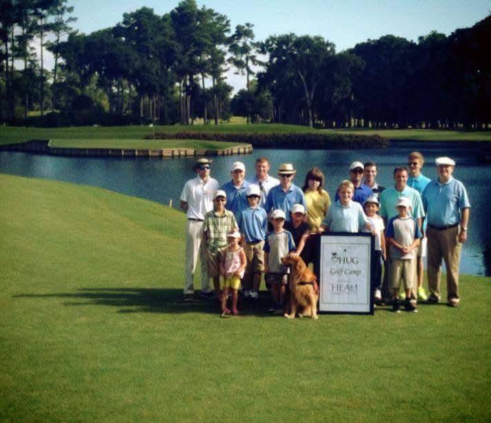 Camp golfers, families and coaches, with a golden retriever, gathered around a HUG Golf Camp and HEAL Foundation sign beside the water at TPC Sawgrass