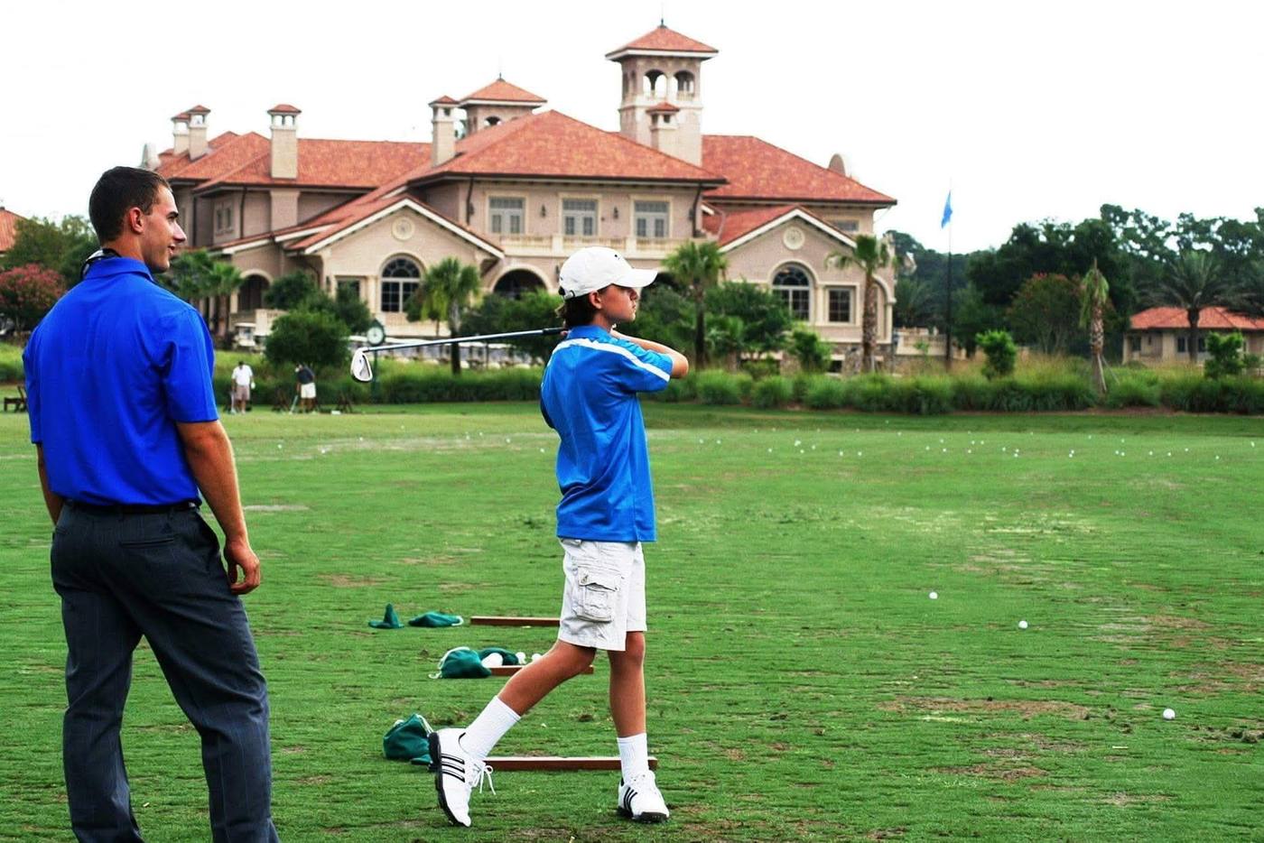 A camp golfer finishes a swing on the range as a coach watches, with the TPC Sawgrass clubhouse behind them