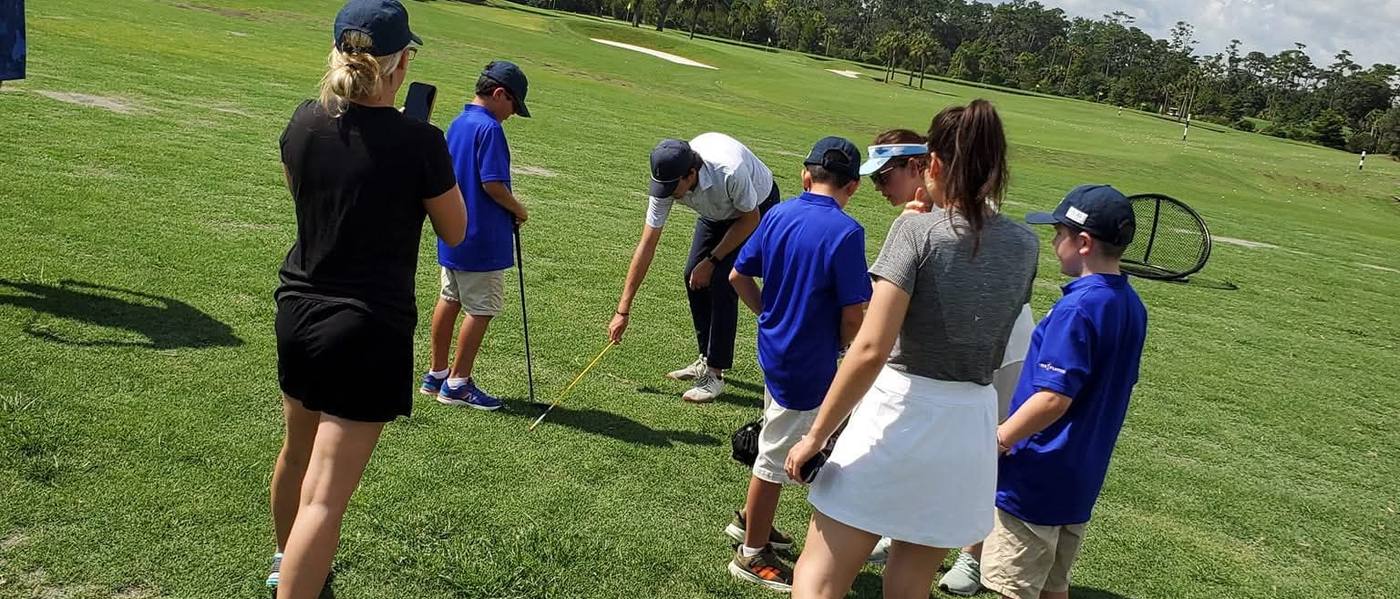 A coach shows camp golfers how to line up a shot while volunteers and parents look on