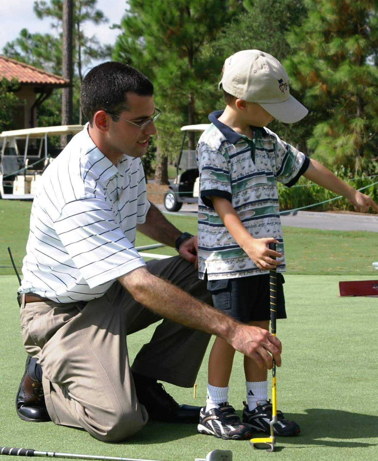 A coach kneels beside a young golfer on the putting green and helps set up a putt